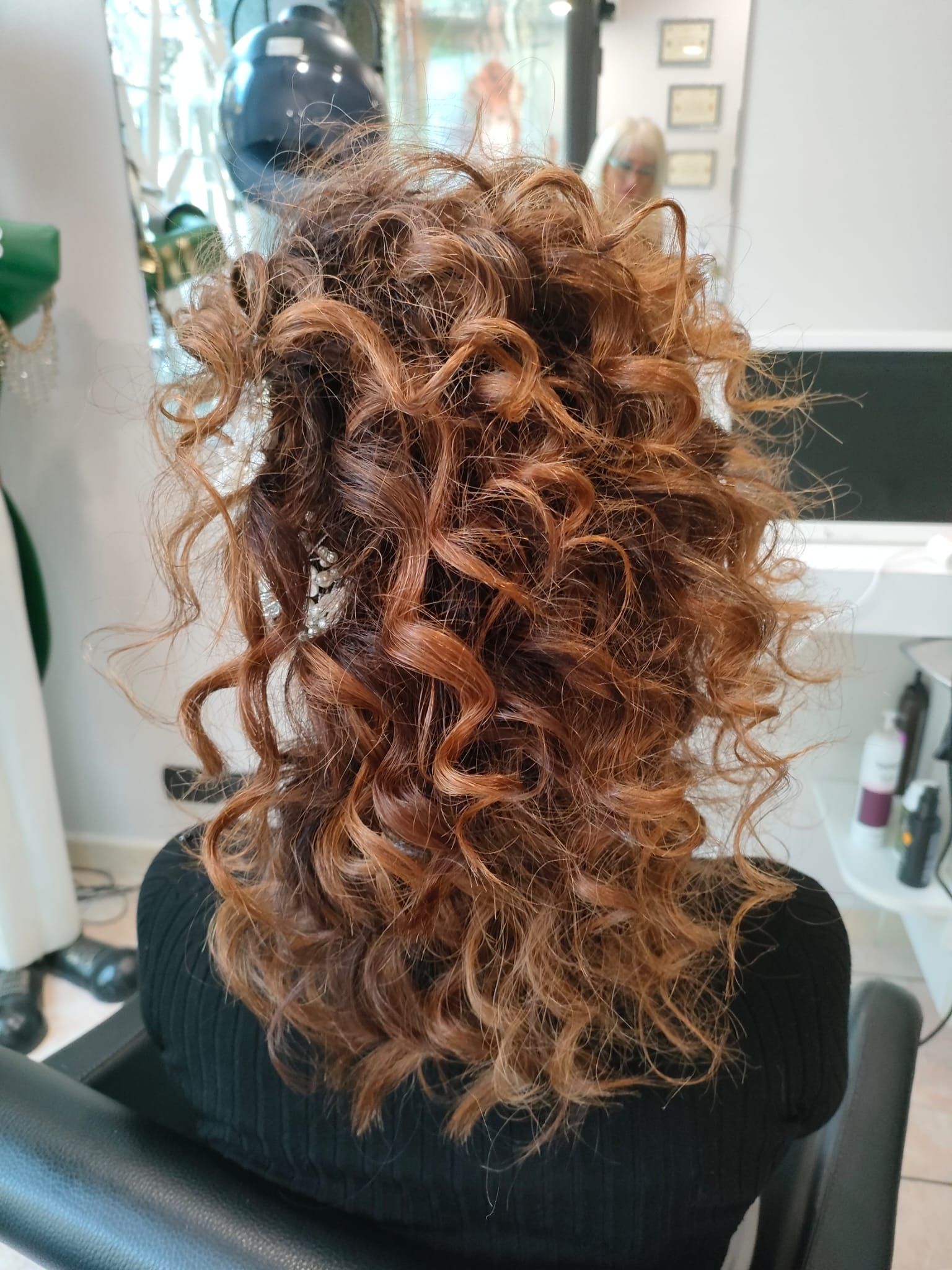 Vista posteriore di una persona seduta su una poltrona da parrucchiere, con capelli ricci, di media lunghezza e voluminosi, di colore castano rossiccio.