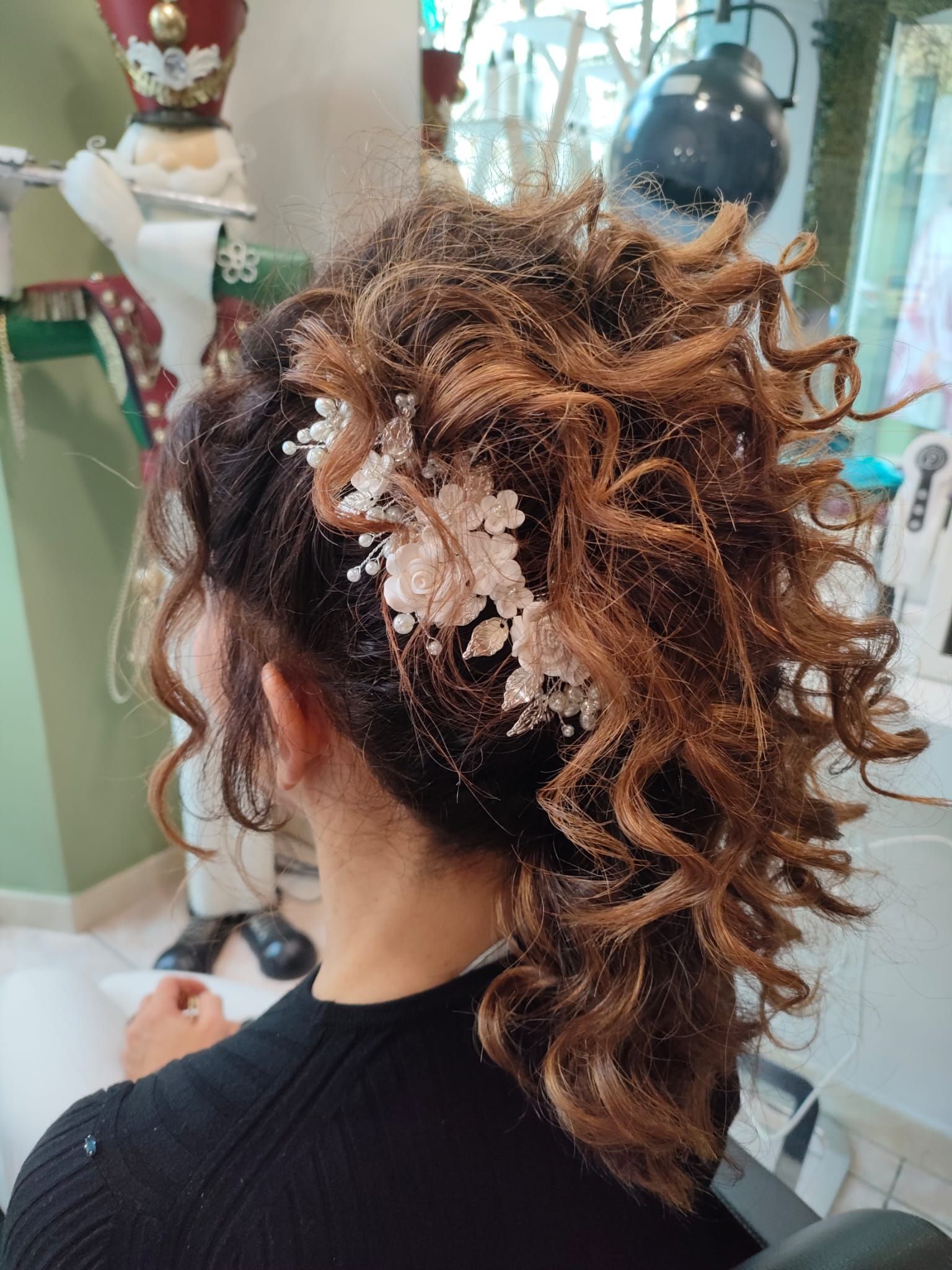 Vista posteriore di una persona con capelli castani ricci raccolti in una mezza coda, adornata da un delicato accessorio floreale bianco.