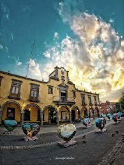 Un edificio amarillo con una torre de reloj y una fila de esculturas de globos terráqueos delante de él.