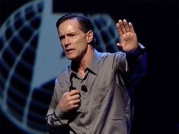 Man speaking on stage, holding a microphone and raising one hand against a blue globe-like backdrop