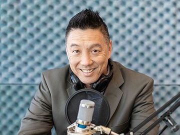 Smiling man in a gray blazer speaking into a microphone in a recording studio