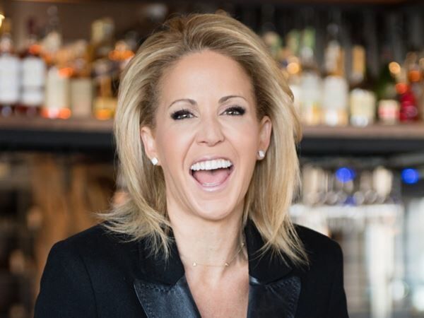 Smiling blonde woman in a black blazer at a bar with blurred bottles in the background