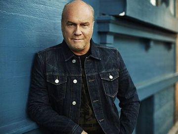 Man in a dark denim jacket leaning against a blue wall, looking at the camera.