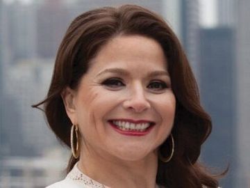 Smiling woman with dark hair and red lipstick, wearing gold hoop earrings, in front of a blurred city skyline