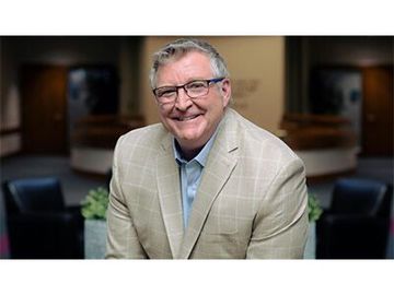 Smiling man in a tan blazer standing in a softly lit office lobby with blurred seating behind him