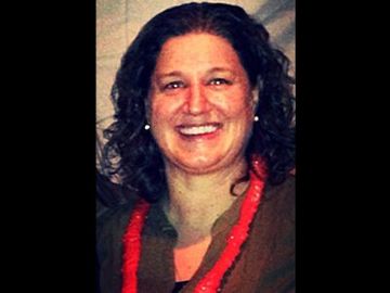 Smiling person with curly dark hair and a red lei against a dark background