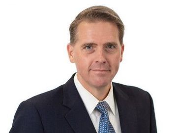 Head-and-shoulders portrait of a man in a dark suit, white shirt, and blue tie on a white background
