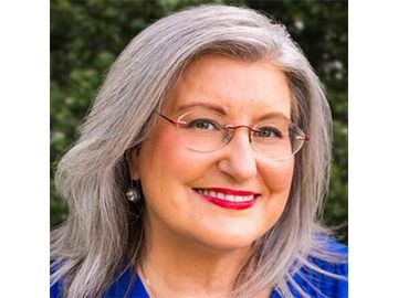 Smiling woman with gray hair and glasses, wearing a blue top against a blurred outdoor background