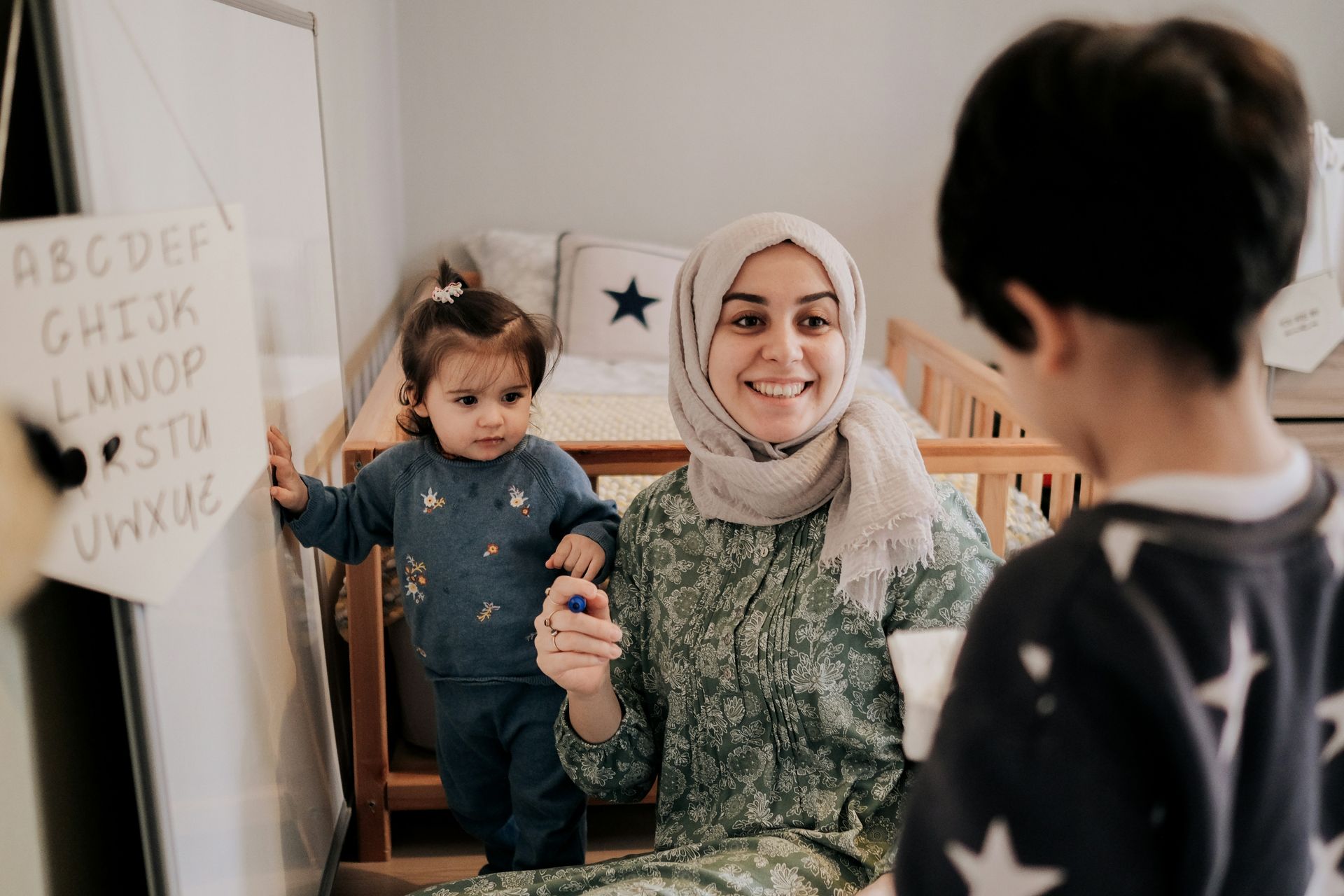 A woman in a hijab teaches two young children. One child looks at her, the other by the whiteboard.