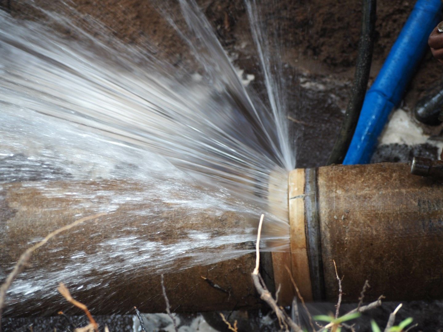 A high-pressure water leak spraying from a joint in a large underground brown pipe.