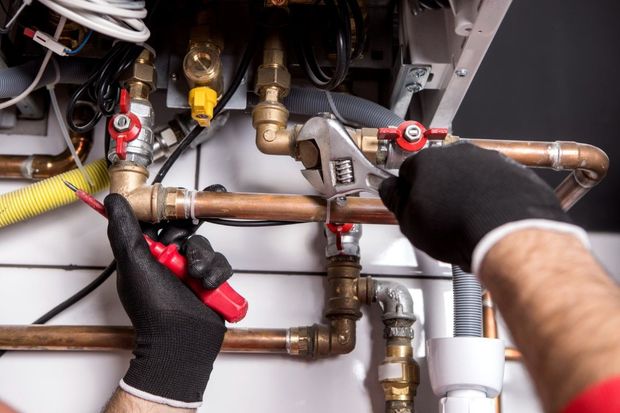 A person wearing black work gloves uses a wrench and a screwdriver to repair copper pipes in a boiler system.