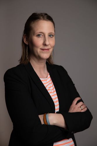 Dorit Horst, bras croisés, vêtue d'un blazer et d'une chemise rayée, souriante. Fond gris.