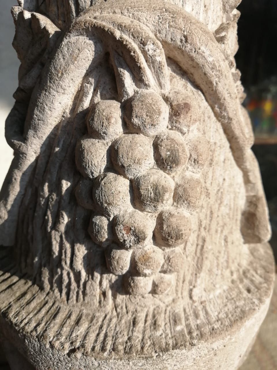 Una escultura en piedra que representa un racimo de uvas rodeado de hojas y detalles de cintas onduladas sobre una base texturizada.