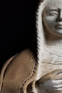 Primer plano de perfil de una escultura de piedra tallada que muestra un rostro sereno y ropa drapeada sobre un fondo negro.