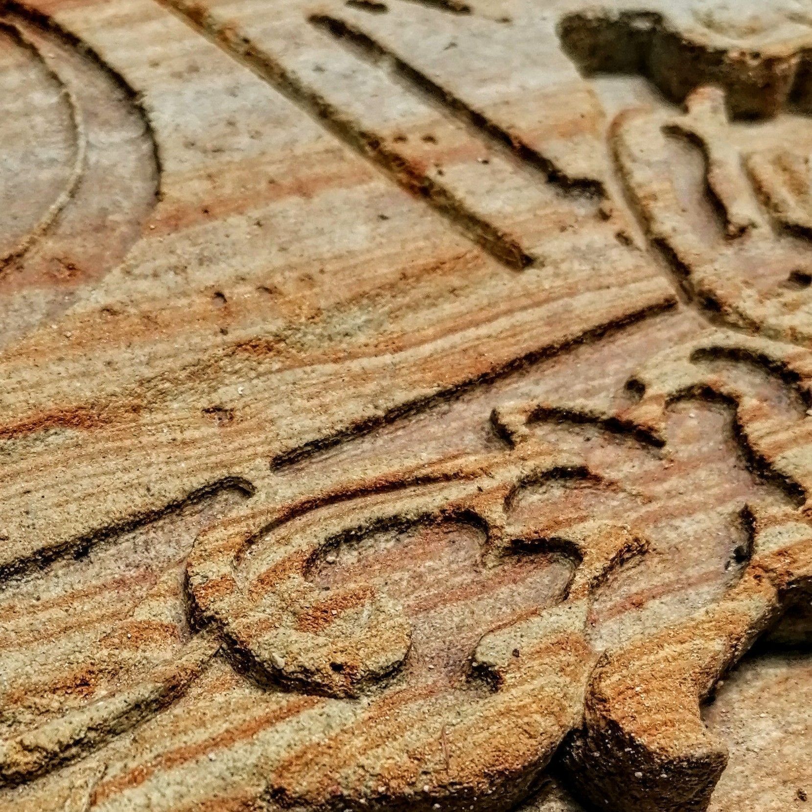 Primer plano de intrincadas tallas decorativas grabadas en una superficie de piedra texturizada de color marrón claro, con la veta de la madera visible.