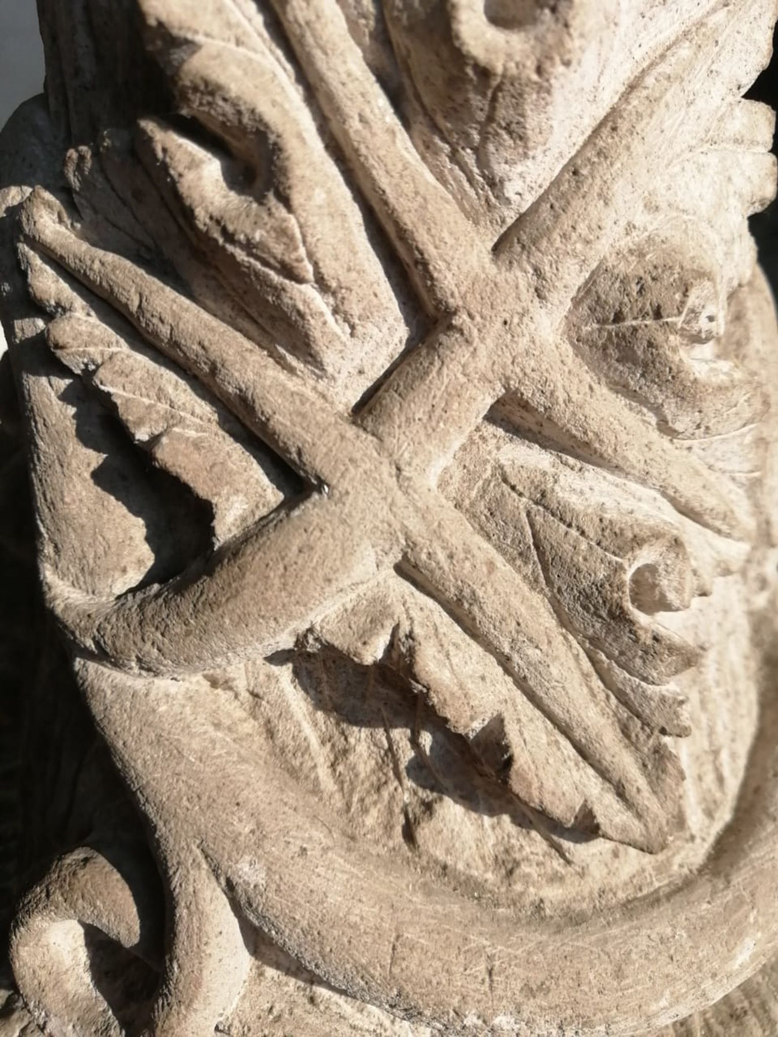 Primer plano de una talla de piedra erosionada que presenta una forma de cruz superpuesta sobre motivos estilizados de hojas de acanto o flores.