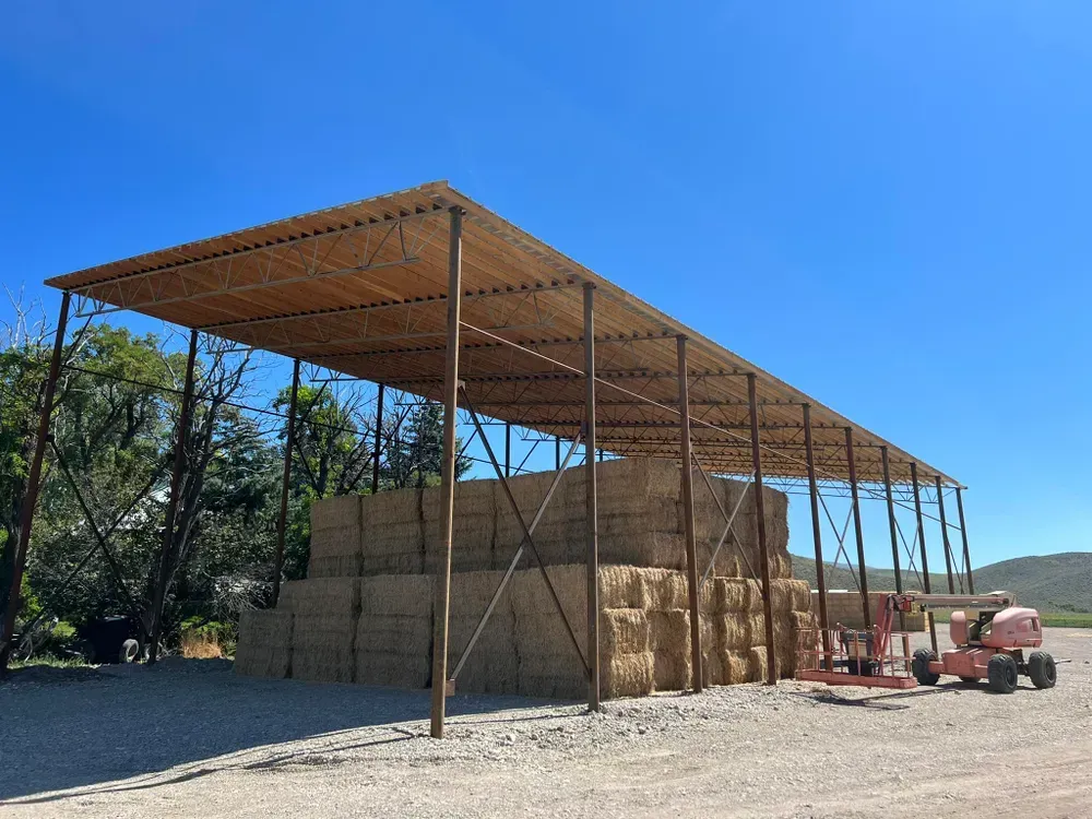 Hay bales stacked under a metal-framed, open-sided shelter on a gravel lot, with a lift and blue sky.