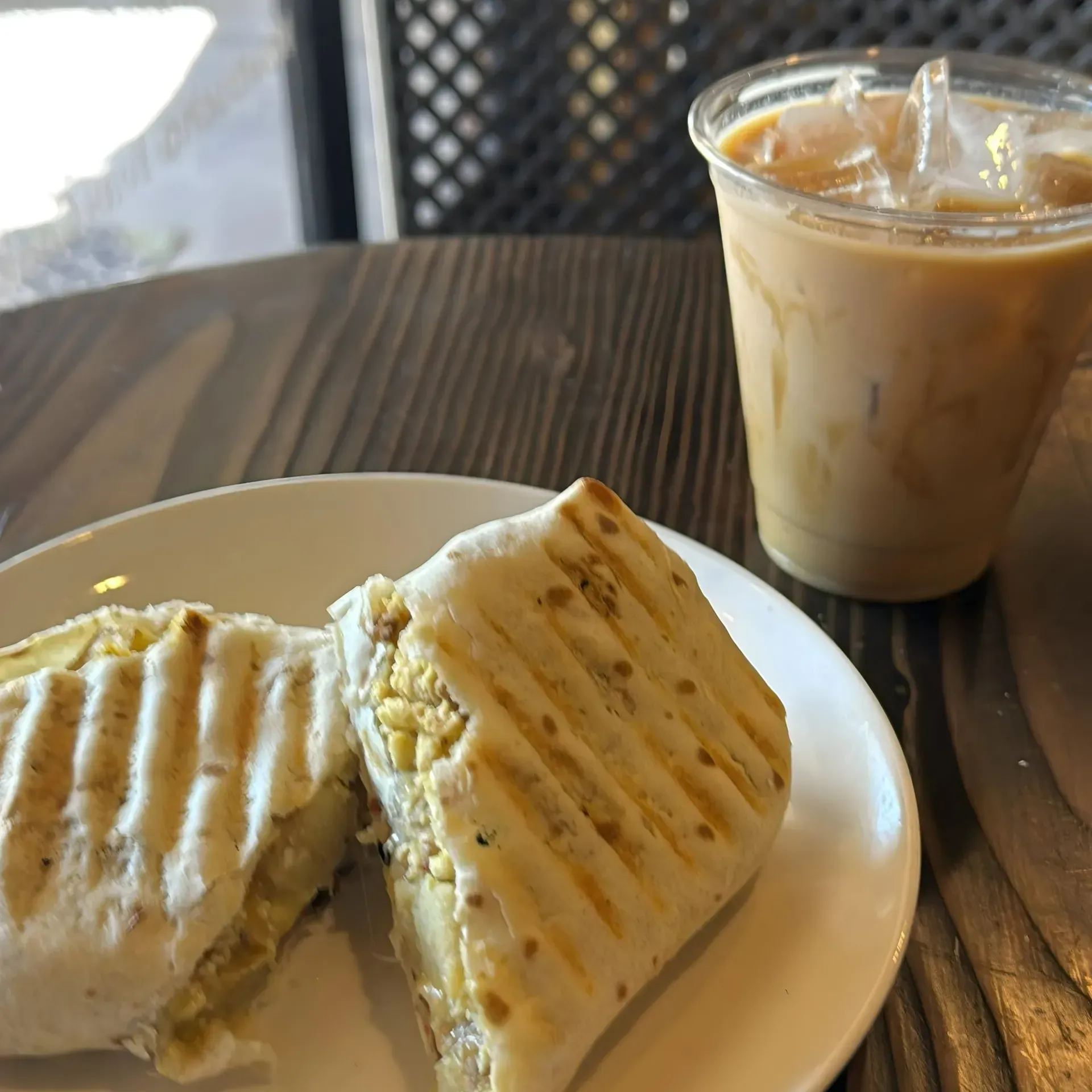 Breakfast sandwich on white plate with iced coffee in plastic cup, on wooden table.