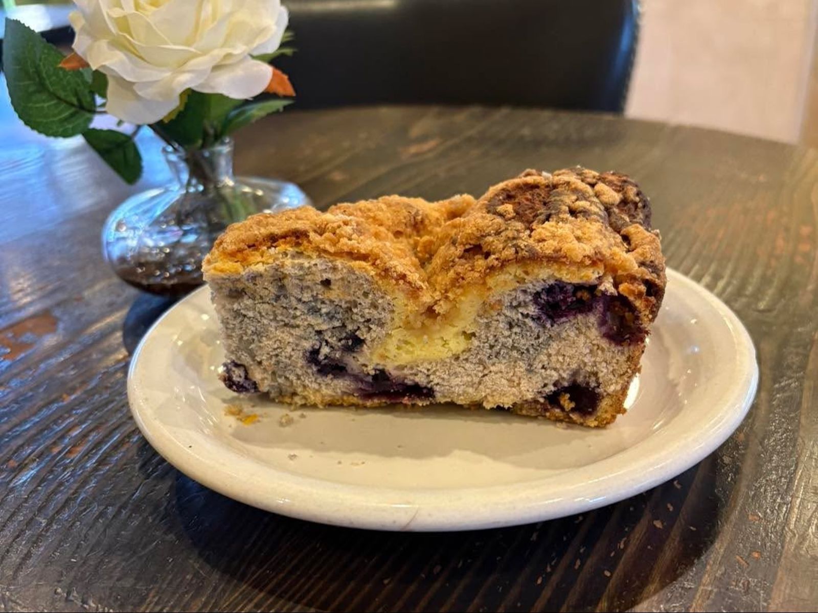 Blueberry coffee cake slice on a white plate with a flower vase on a dark wood table.