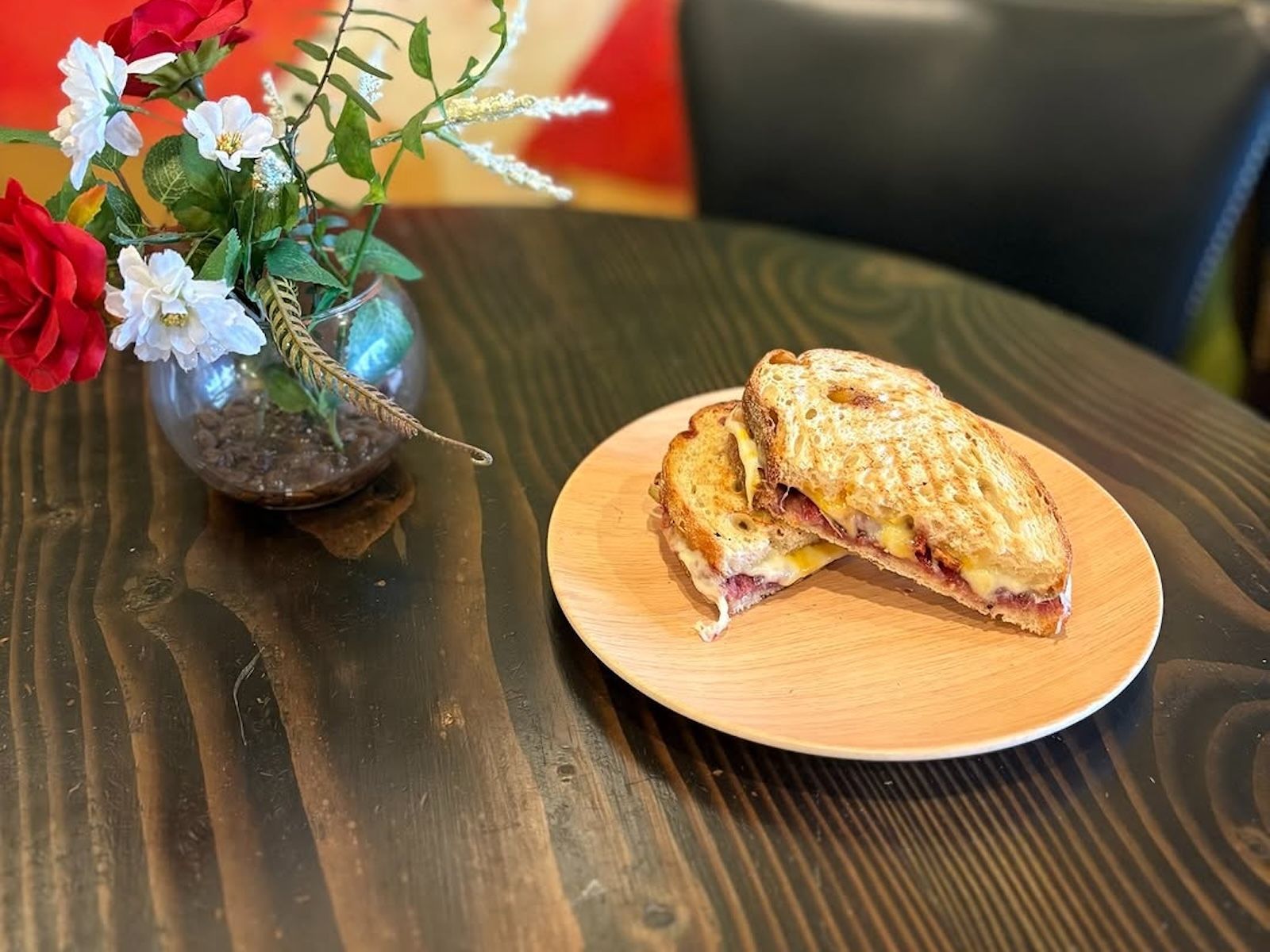 Grilled sandwich on a wooden plate next to a vase of flowers on a dark wood table.