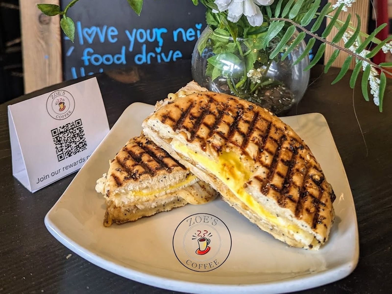 Grilled cheese sandwich on a plate with food drive sign and flowers.