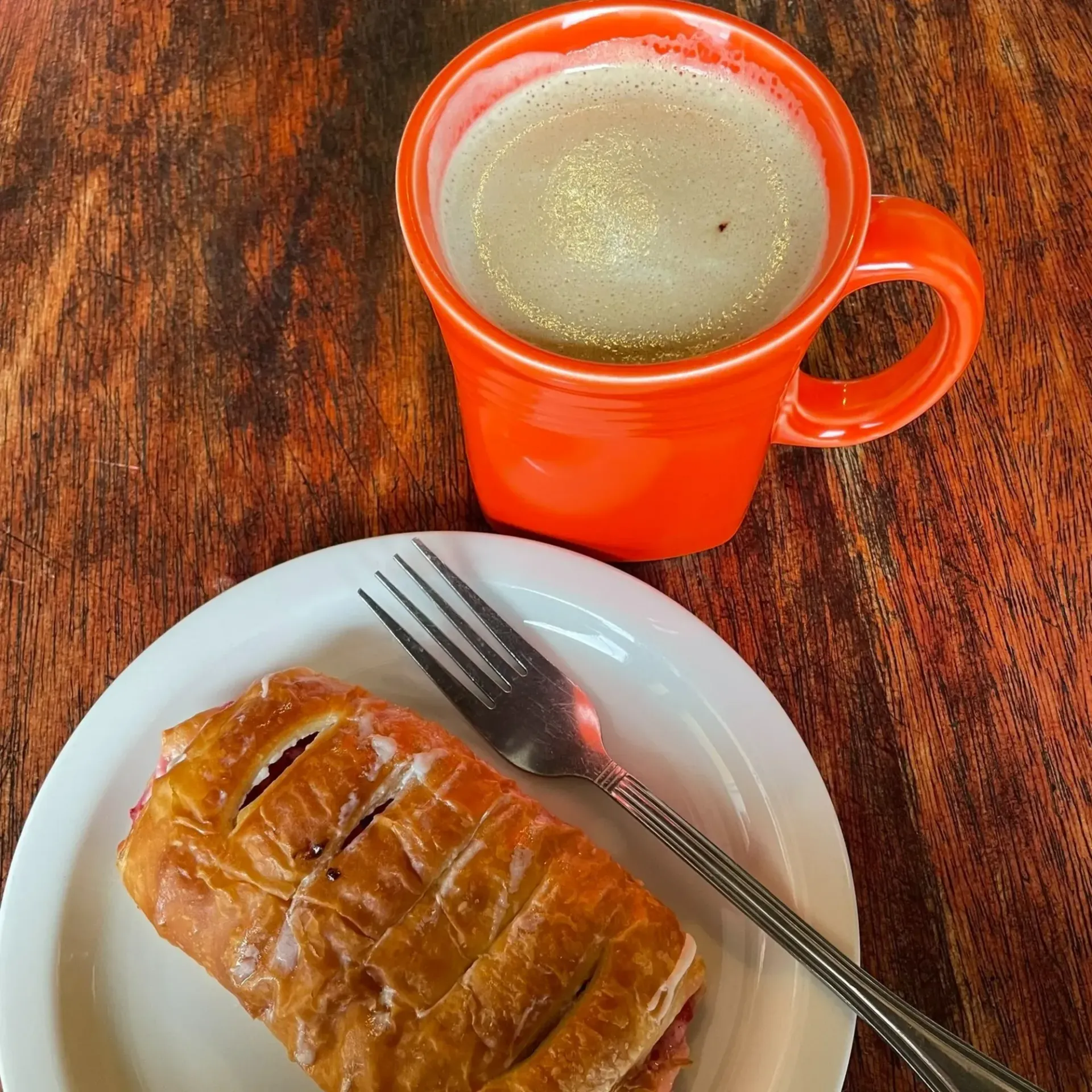 Orange mug with foamy drink next to pastry on a white plate with a fork, on a wood table.