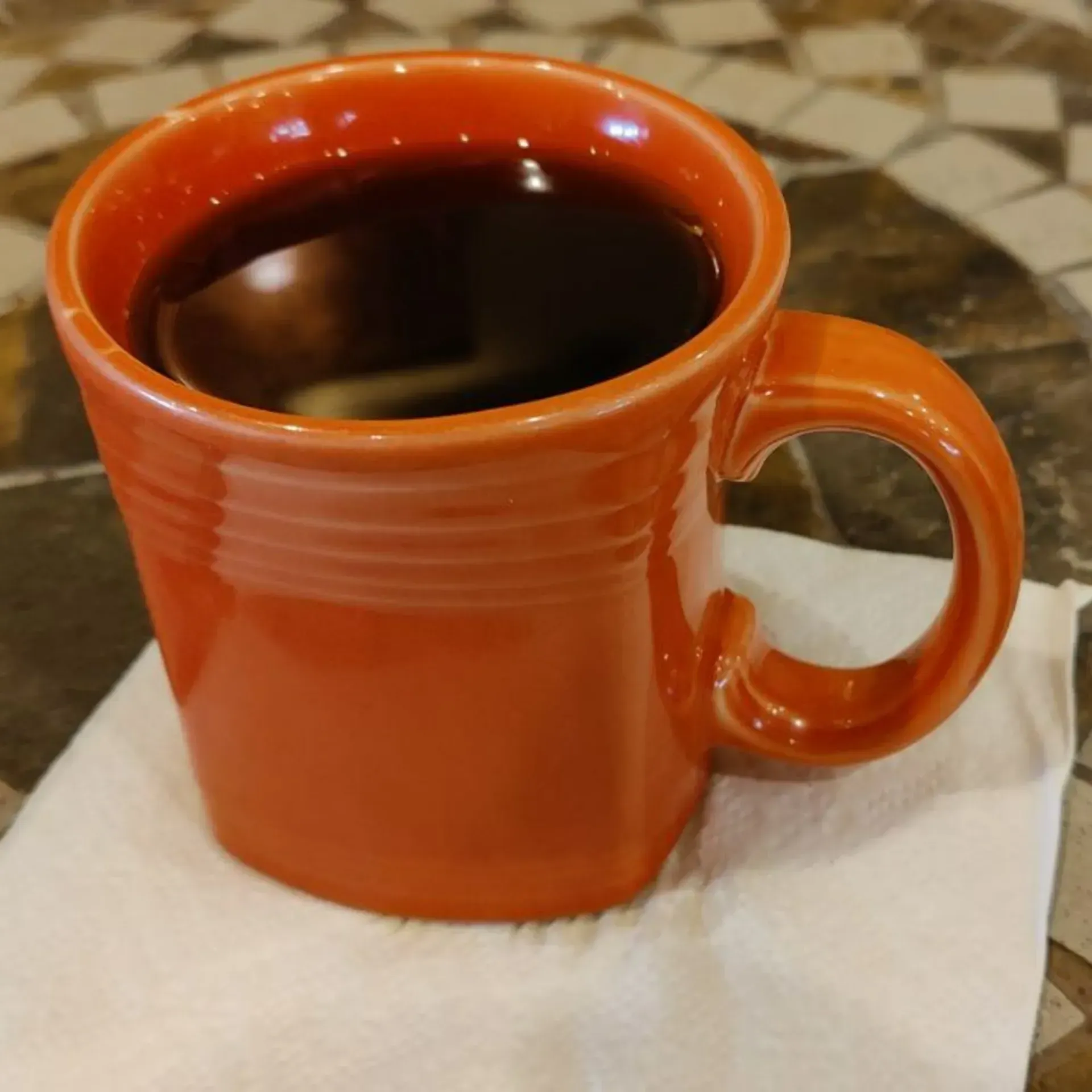 Orange mug filled with dark liquid, on a white napkin, on a patterned surface.