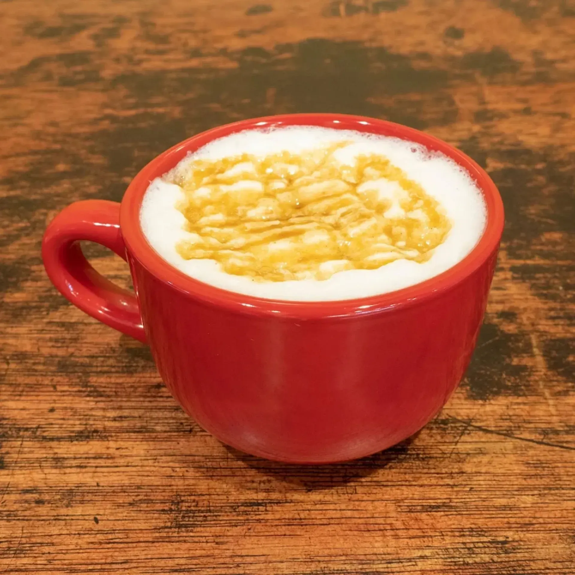 Red mug of coffee with foamy milk and caramel drizzle on a wooden surface.