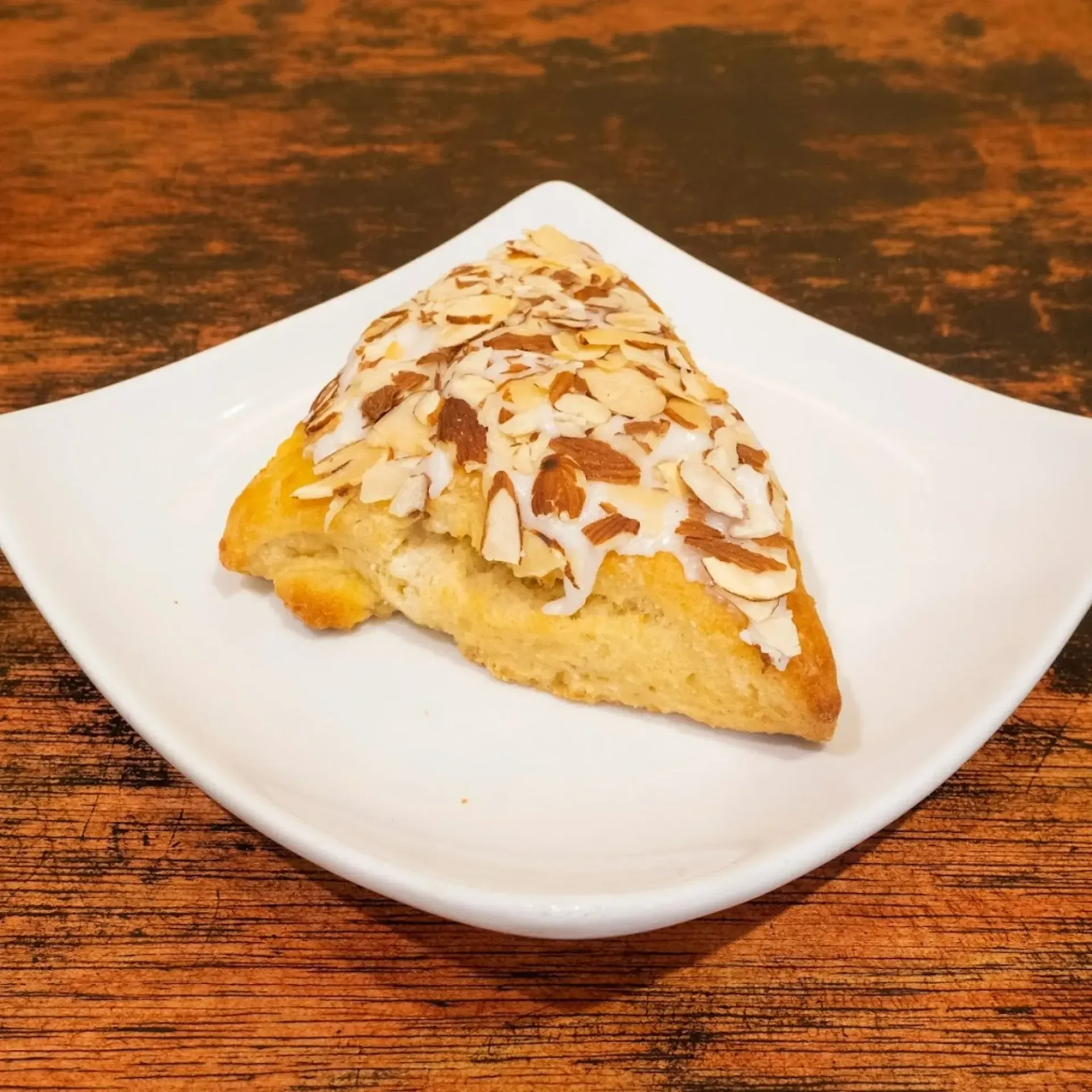 Scone with almond glaze and slivered almonds on white square plate, set on wooden table.