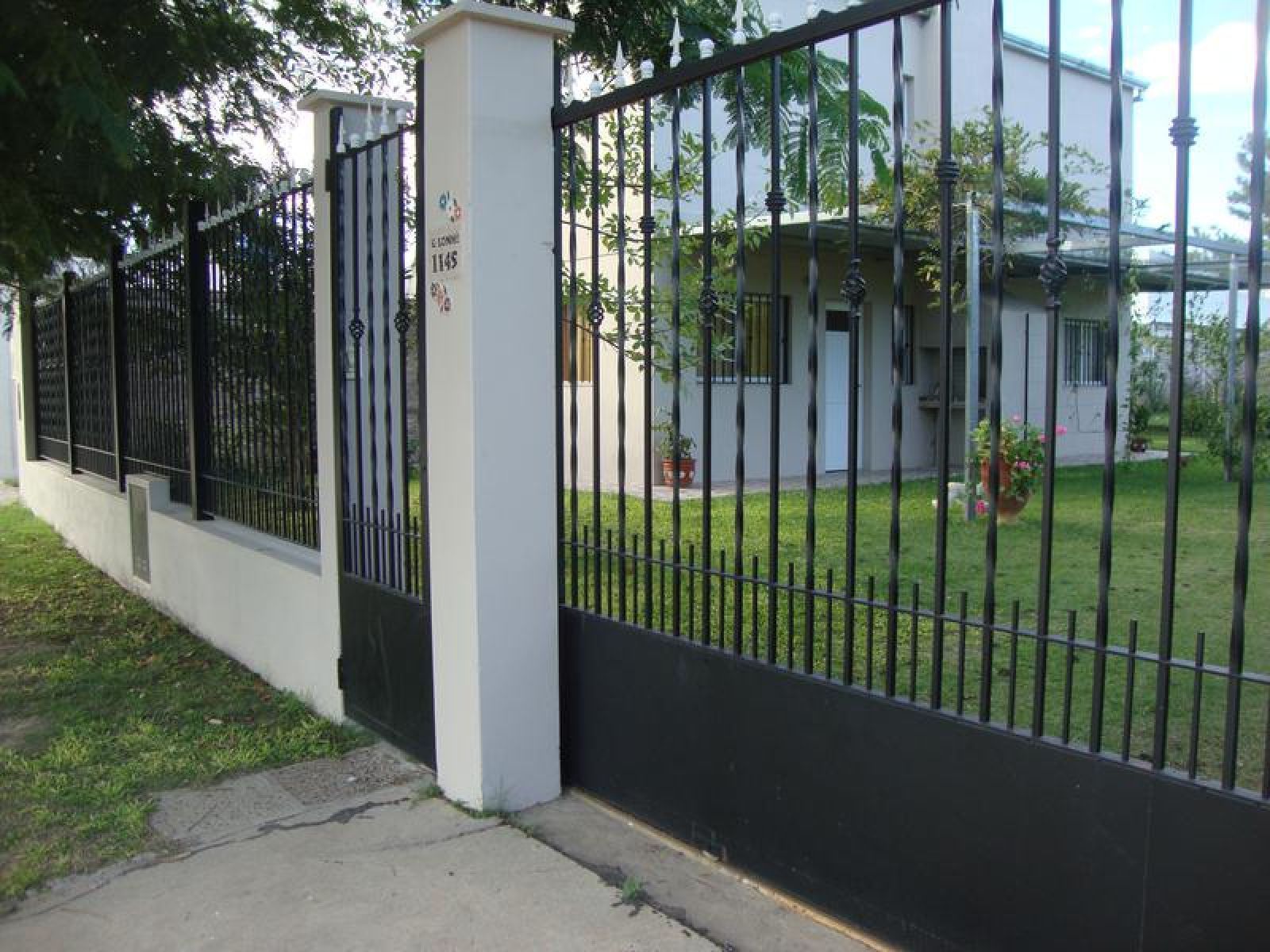 Verja de hierro forjado negro con puerta de columnas blancas, frente a una casa de color claro.
