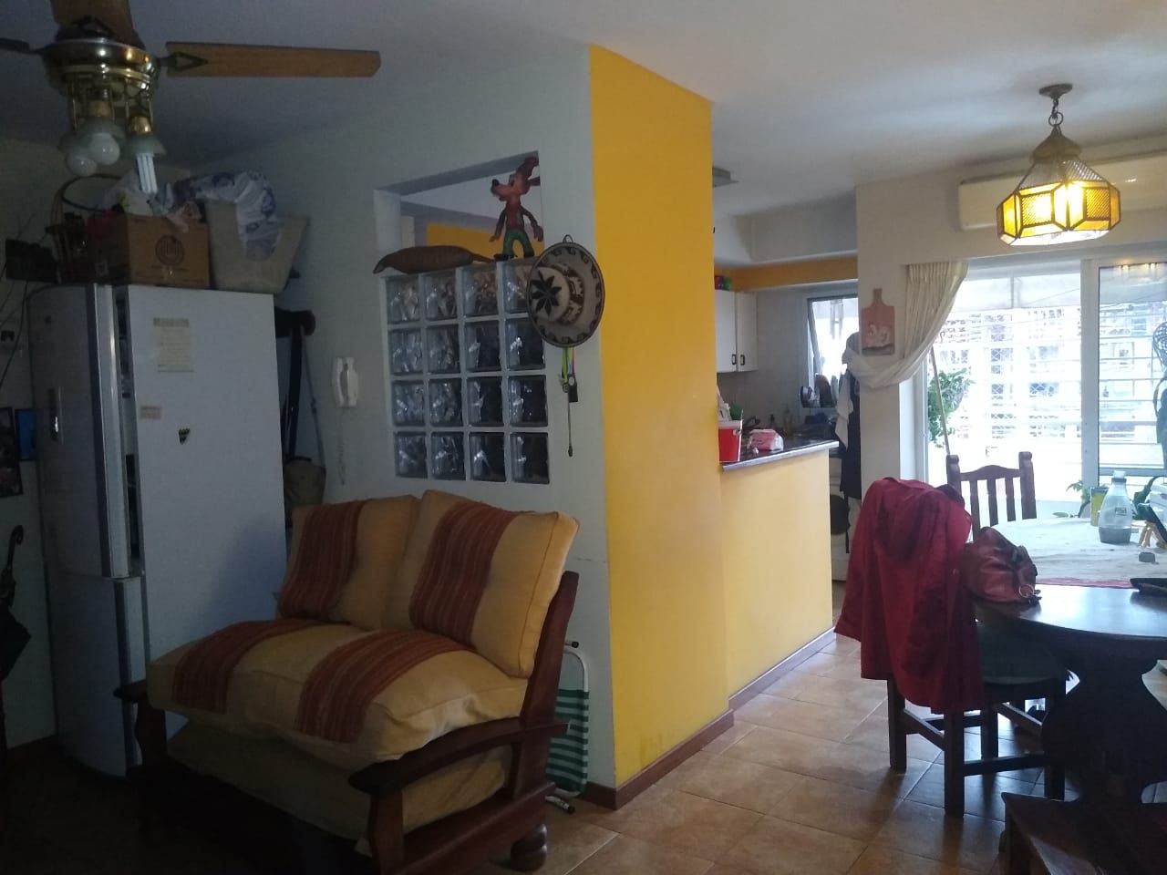 Sala de estar con pared de acento amarillo, cocina al fondo, silla y mesa.