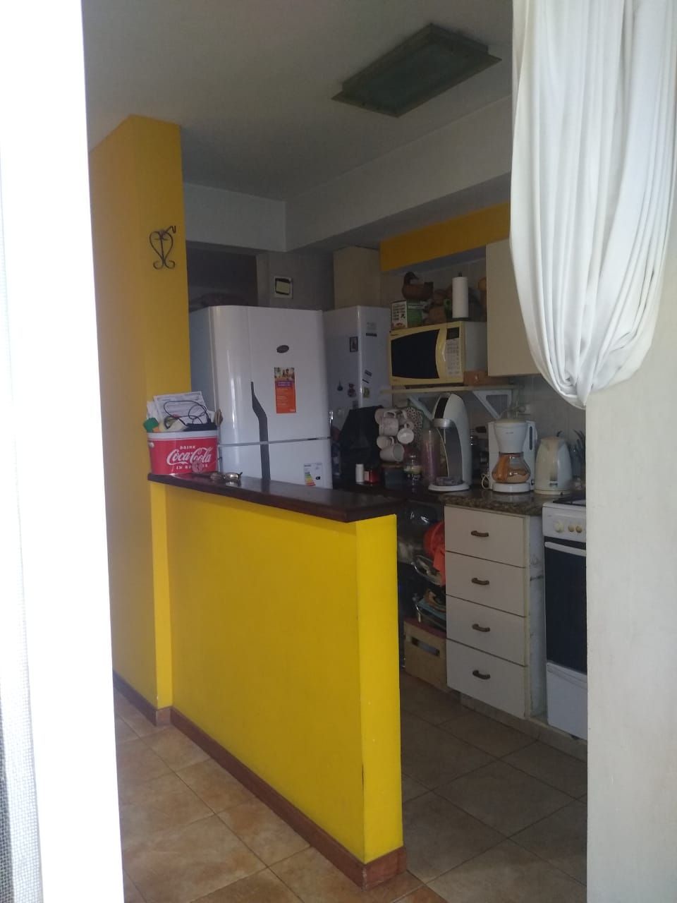 Sala de estar con pared de acento amarillo, cocina al fondo, silla y mesa.