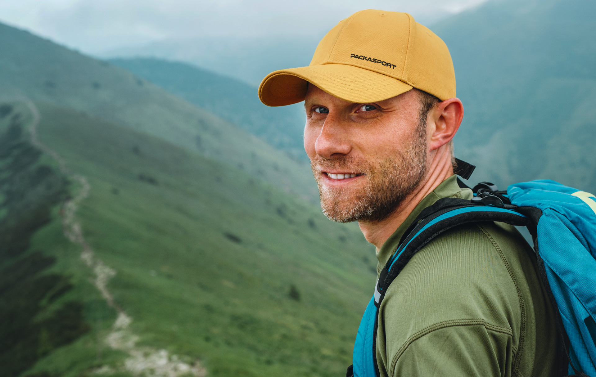 A man wearing a yellow hat and a blue backpack is standing on top of a mountain.