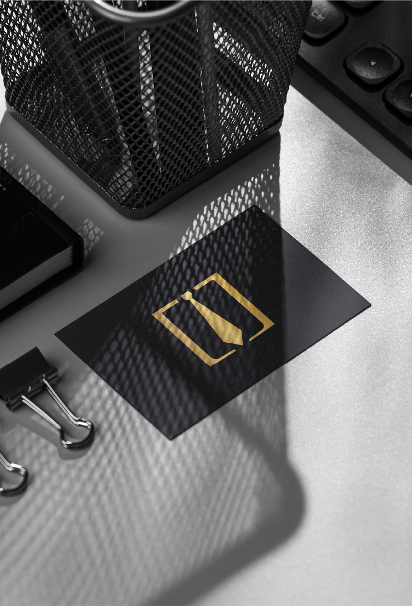 A business card with a gold tie icon inside a gold square border, resting on a desk with office supplies.