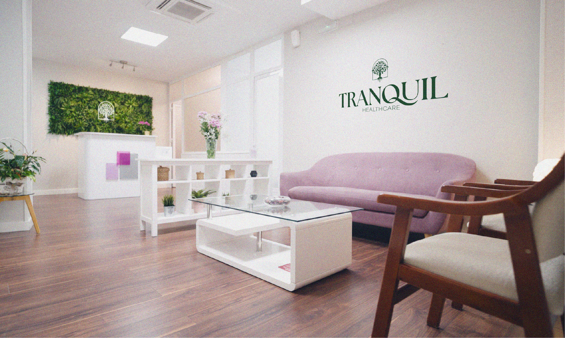 A modern, brightly lit waiting room featuring a pink sofa, glass coffee table, and a reception desk with a moss wall.