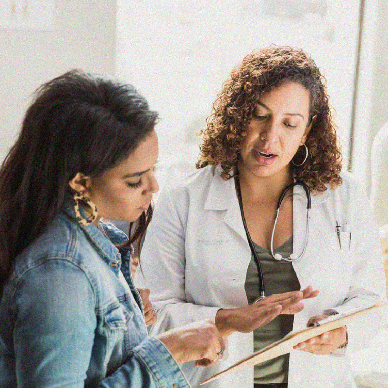 A healthcare provider in a white lab coat holds a clipboard and talks to a patient, who is gesturing toward the document.