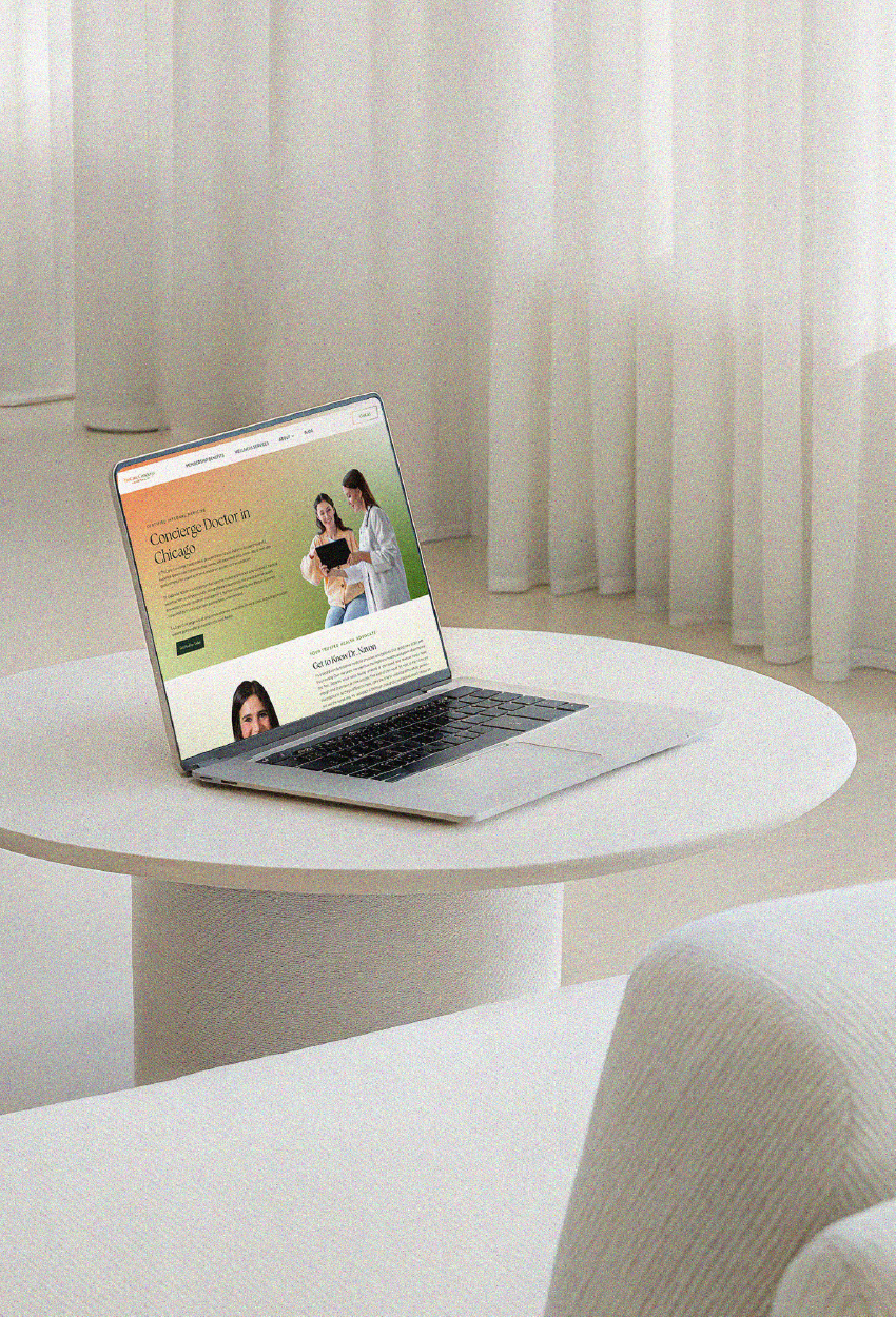 A laptop sits on a round white table in a bright room with sheer curtains, displaying a website with a nature-themed hero.