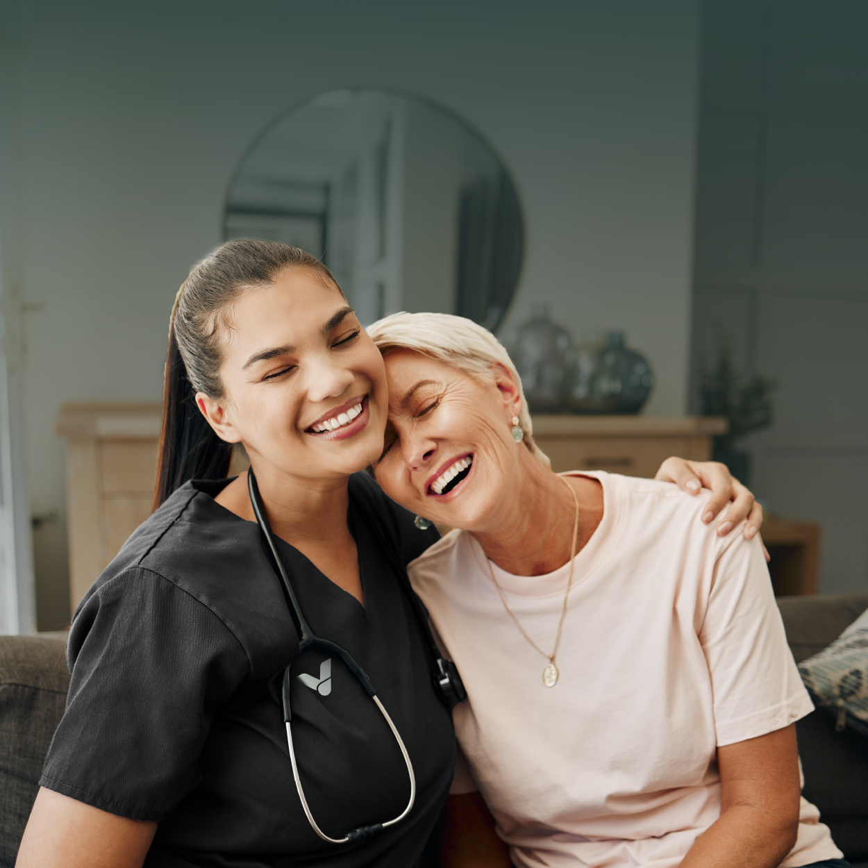 A person in dark scrubs with a stethoscope around their neck hugs another person, both smiling with eyes closed.