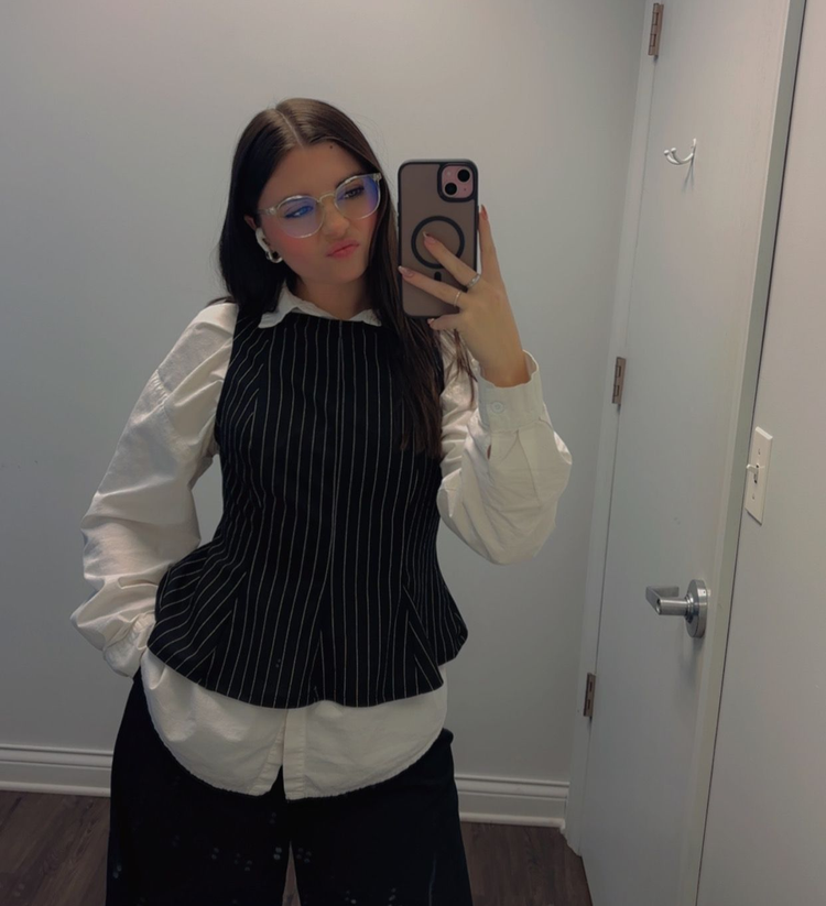 A person wearing a pinstripe vest over a white button-down shirt and glasses, taking a mirror selfie in a room.