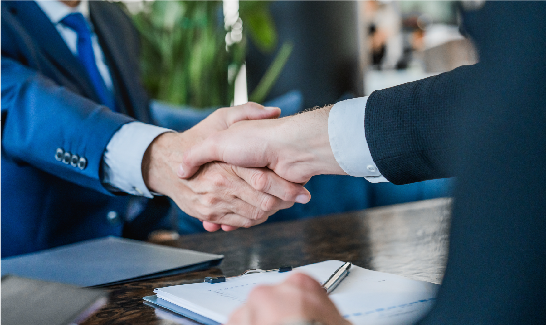 Two people in business suits shake hands over a desk with a notebook and pen during a professional meeting.