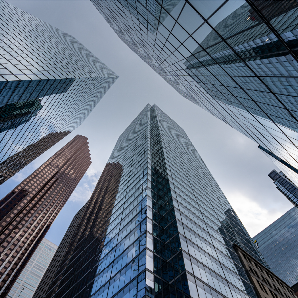 A low-angle view of multiple modern glass skyscrapers reaching toward a bright, cloudy sky in a dense urban center.
