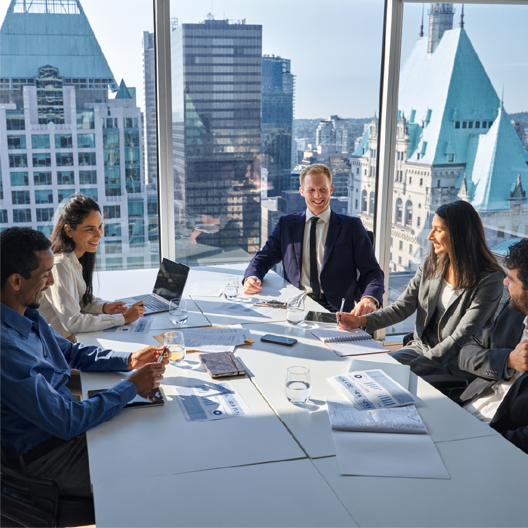 A group of professionals sit around a table for a meeting in a modern office with large windows overlooking a city.