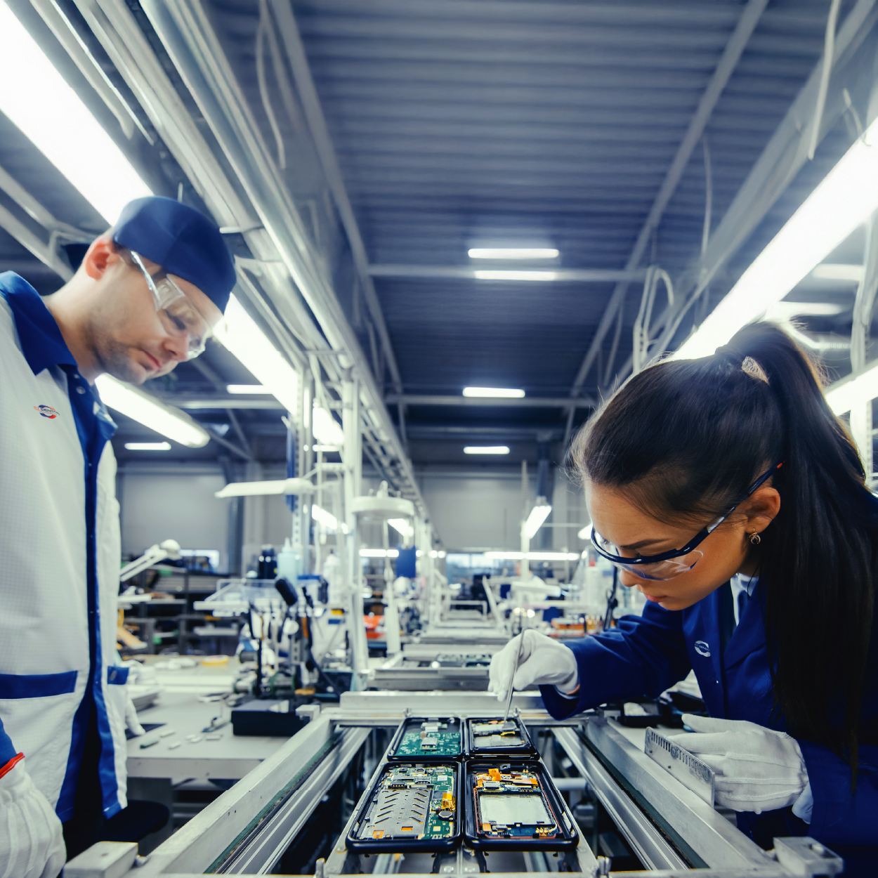 Two technicians in safety glasses and work coats assemble electronic components on a factory production line.