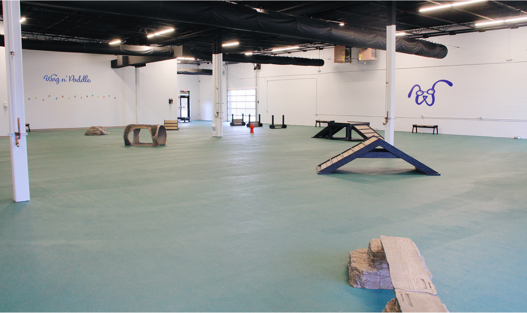 Indoor dog training facility with agility equipment, including ramps, tunnels, and hurdles on a large, green rubber floor.