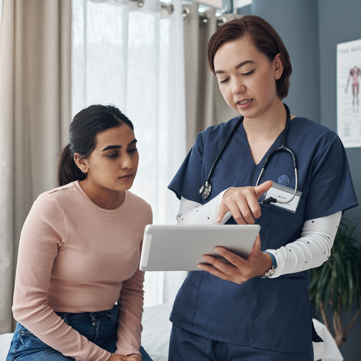 A healthcare provider in blue scrubs points at a tablet screen while consulting with a patient in a medical office.