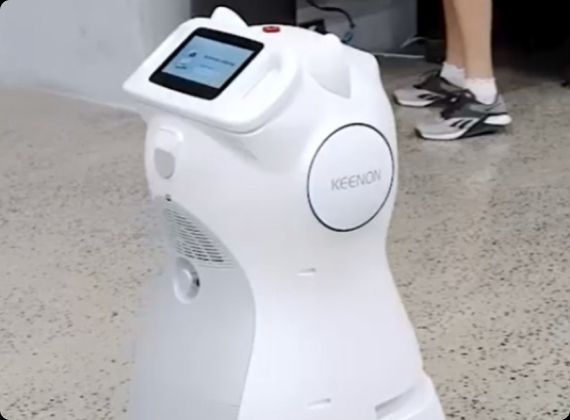 White robot with a screen, indoors. Person in sneakers in the background.