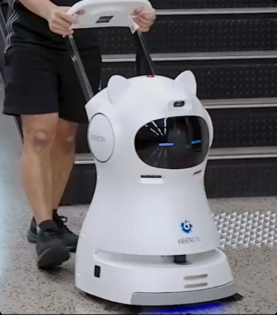 Person pushing a white Keelson cleaning robot with cat-like ears; near stairs.