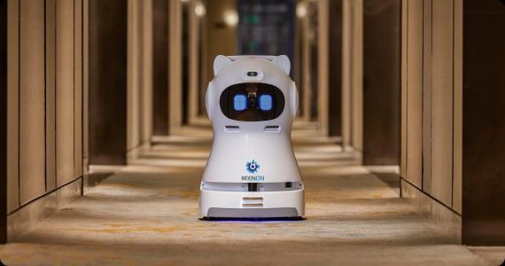 White delivery robot in hotel hallway with blue-lit eyes and logo, centered on the floor.
