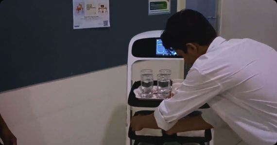 A person places items from a robot tray; glasses of water and towels. Hospital setting, robot white.