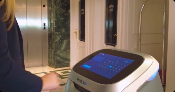 Robot with touch screen next to a person in an elevator lobby.